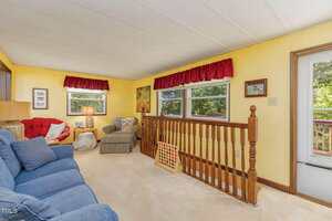 A cozy living room with yellow walls, beige carpet, blue sofa, two armchairs, a wooden railing, large windows with red valances, a lamp, framed pictures, and a door leading outside.