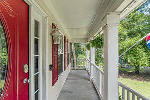 A covered front porch with a red door, white columns, a hanging lantern, potted fern, and railing, overlooking a grassy yard and trees. A flag is partially visible on the right.