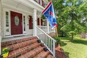 Brick steps with white railings lead to a porch with a red door and sidelights. A blue sports flag hangs by the entrance. Potted plants line the steps, and a green lawn with trees is visible in the background.