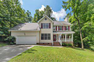 Charming two-story house in Creedmoor with light-colored siding, red shutters, an attached double garage, and a covered front porch with columns on 2750 Michelle Court. Sloped lawn is surrounded by trees under a partly cloudy sky.