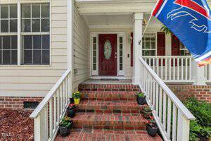 A brick porch with four steps leads to a red front door with glass panels on a cream-colored house. There are five potted plants on the steps and a blue and red flag hanging on the right side.