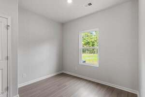An empty, small room with light gray walls, a white ceiling, a single window showing a green outdoor scene, wood-style flooring, a closed white door, and a white vent near the ceiling.