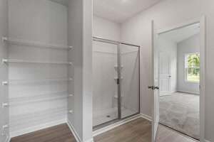 A bathroom with an empty white wire shelving unit, a glass-enclosed shower with sliding doors, light wood flooring, and an open door leading to a carpeted room with a window.
