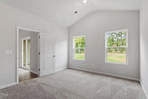 A bright, empty room with light gray carpet, white walls, two windows showing greenery outside, a white door, and an open doorway leading to a space with wood flooring. Ceiling has a recessed light and vent.