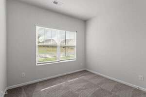 A small, empty room with light gray walls, a beige carpeted floor, and a window with white blinds overlooking a suburban street and lawns.