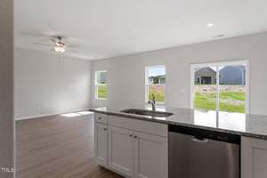 A modern kitchen with white cabinets, a granite countertop, and a stainless steel dishwasher, overlooking a living area with large windows and light wood flooring. The space is empty and well-lit with natural light.