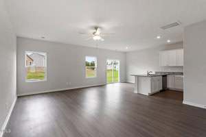 A spacious empty living area with light gray walls, dark wood flooring, a ceiling fan, and three windows. An open kitchen with white cabinets and granite countertops is visible in the corner. A door leads outside.