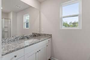 A modern bathroom with a white vanity, granite countertop, under-mount sink, two windows, and a partial view of a glass shower enclosure reflected in the large wall mirror.