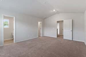 Empty carpeted bedroom with white walls, two open doors leading to a bathroom and closet, and a closed door on the right. Natural light enters through a small window near the bathroom. Ceiling lights are on.