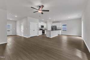 Open-concept living area with light gray walls, wood-style flooring, a ceiling fan, and recessed lighting. The space includes a modern kitchen with white cabinets, stainless steel appliances, and an island with a sink.