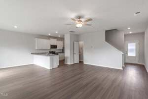 Unfurnished open-plan living area with light wood flooring, white walls, and ceiling fan; kitchen in the background with white cabinets and stainless steel appliances; staircase and back door visible.