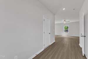 A view of an empty room with light-colored walls, wood flooring, two closed white doors on the left and right, a window at the far end, and a ceiling fan with light in the background.