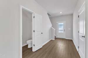 A hallway with light gray walls and wood flooring features a white door leading to a small bathroom with a toilet, a staircase, a back door with frosted glass, and another closed door on the right.