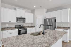 Modern kitchen with white cabinets, stainless steel appliances including a refrigerator, oven, and microwave, granite countertops, and a large island with a sink and a black faucet. Walls are light gray.