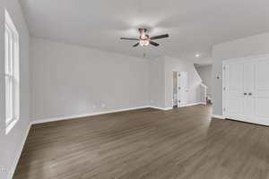 Spacious, empty room with light gray walls, wood-style flooring, a ceiling fan with lights, double doors on the right, and a staircase leading up at the back. Natural light enters from large windows on the left.