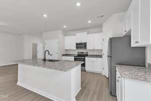 Modern kitchen with white cabinets, stainless steel appliances, granite countertops, a large central island with a black faucet, light wood flooring, and recessed ceiling lights. Walls are painted a light neutral color.