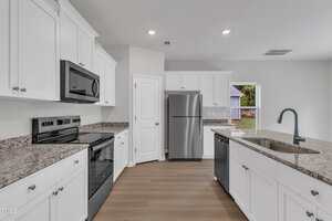 Modern kitchen with white cabinets, granite countertops, stainless steel refrigerator, black oven and microwave, dishwasher, and a sink on an island. There is a door and window letting in natural light.
