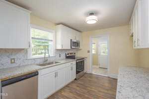 Modern kitchen with white cabinets, stainless steel appliances, granite countertops, and a herringbone-patterned backsplash. There is a window above the sink and wood-style flooring throughout the room.