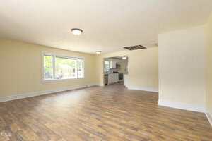 A spacious, empty living room with light yellow walls, wood-style flooring, two ceiling lights, a large window, and an open view into a kitchen with white cabinetry and appliances.