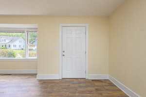 A white door is centered on a light yellow wall, with a window to the left showing houses and greenery outside. The room features light brown wood flooring and white trim.