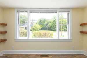A large three-panel window with white trim looks out onto a green yard with bushes and trees. The interior room has cream-colored walls, wooden floors, and built-in wooden shelves on both sides of the window.