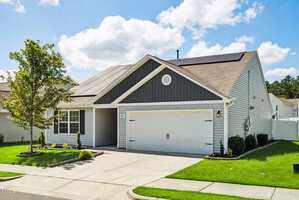 A single-story house with gray siding, white trim, a double garage, and solar panels on the roof. The front yard has green grass, small shrubs, a tree, and a concrete driveway under a partly cloudy sky.