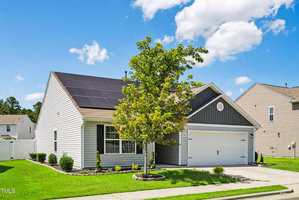 Single-story house with gray siding, white trim, and solar panels on the roof. A tree partially obscures the front, with a driveway leading to a two-car garage. The sky is blue with scattered clouds.