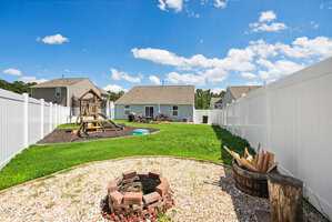 A backyard with white vinyl fencing, a playset with slides, a patch of green grass, a circular fire pit lined with bricks, gravel surrounding the fire pit, and a wooden barrel filled with firewood under a blue sky.