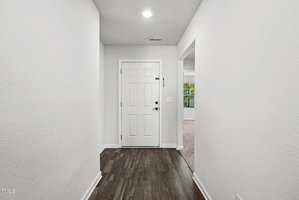 A hallway with dark wood flooring leads to a white front door. The walls are white, and a doorway on the right opens to a carpeted room with a window. A ceiling light is above the entryway.