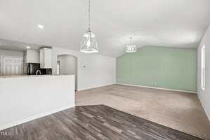 Open-concept living space with dark wood flooring leading to carpeted area, green accent wall, two geometric pendant lights, and a kitchen with white cabinets and granite countertops visible through an arched opening.