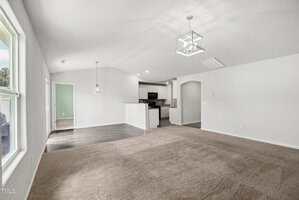 A bright, unfurnished open-concept living area with beige carpet, white walls, and a view into the kitchen and dining space. The kitchen has dark flooring, white cabinets, and modern light fixtures.