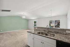 A view from the kitchen showing a double sink with granite countertops, white cabinets, and an open living area with beige carpet, a green accent wall, and windows letting in natural light.