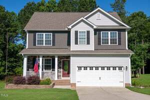 Charming two-story suburban home at 933 Weatherby Lane, Creedmoor, with white siding, gray accents, double garage, front porch adorned with an American flag, and a neatly landscaped yard. Trees line the background.