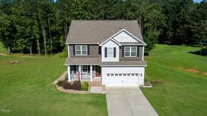 A two-story house with a covered front porch, attached two-car garage, and driveway, surrounded by a large green lawn with trees in the background.
