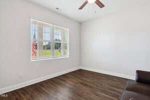 A mostly empty room with white walls, a large window with blinds, wood flooring, a ceiling fan with a light, and a small part of a brown couch visible in the lower right corner. An American flag is seen outside the window.