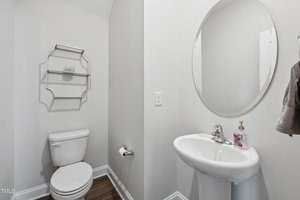 A bathroom with a white toilet, a wall-mounted metal shelf, a white pedestal sink with a round mirror above it, a towel hanging on the wall, and a soap dispenser on the sink. The floor is dark wood.