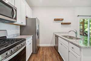 A modern kitchen with white cabinets, stainless steel appliances, granite countertops, a gas stove, wood floors, two wooden floating shelves, and a large window overlooking a green outdoor area.