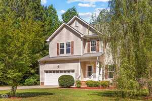Two-story beige house with white trim, brown shutters, and a front porch. The house has a two-car garage and is surrounded by green trees and grass under a blue sky.