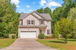 Charming two-story beige house with white trim at 2555 Winding Creek Lane, Franklinton. Features a two-car garage, concrete driveway, lush front yard with shrubs and small trees, and wooded areas in the background under a partly cloudy sky.