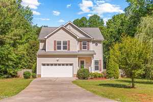 Charming two-story beige house with white trim at 2555 Winding Creek Lane, Franklinton. Features a two-car garage, concrete driveway, lush front yard with shrubs and small trees, and wooded areas in the background under a partly cloudy sky.