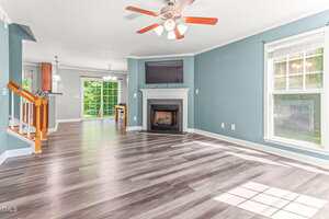 A spacious living room with light gray wood flooring, teal walls, a white-framed window, ceiling fan, and a fireplace below a mounted TV. Dining area and kitchen with large windows are visible in the background.