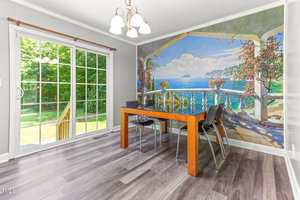 A dining area with a rectangular wooden table and four black chairs sits on gray wood-look flooring. A mural of a seaside balcony scene covers one wall, and large glass doors open to a green yard outside.