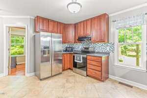 Modern kitchen with stainless steel refrigerator, oven, and microwave, brown cabinets, tile floor, and a window with a view of green trees. Doorway leads to another room with wood flooring.