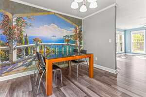 A small dining area with a wooden table and four black chairs is set against a large wall mural depicting a scenic ocean view from a terrace. The room has gray walls and light-colored wood flooring.