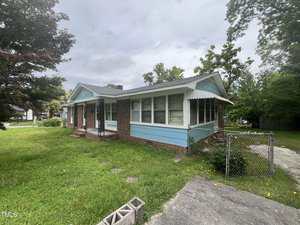 Single-story brick and blue-sided house with a covered front porch, enclosed sunroom, and chain-link fence. The yard is grassy with some bushes and trees. The sky is overcast.