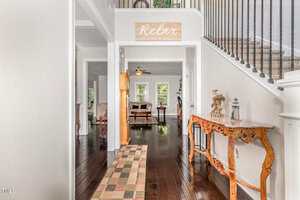 A hallway with dark hardwood floors, a patterned rug, a wooden console table, and a staircase with black railings. A sign above a doorway reads "Relax, Unwind & Enjoy." Living areas are visible beyond the hallway.