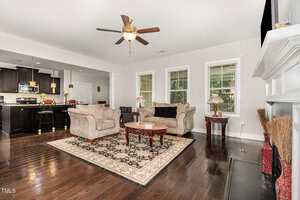 A living room with dark wood floors, beige sofas, a patterned area rug, a coffee table, and large windows. The kitchen with dark cabinets and a breakfast bar is visible in the background.