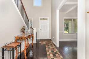 A bright entryway with a white front door, a window above it, a wooden console table with decorative items on the left, a patterned rug on dark wood floors, and an open view into an adjacent room.