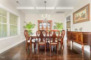 A dining room with a wooden table and six chairs, a glass chandelier above, a china cabinet in the back, two large potted plants, a sideboard on the right, hardwood floors, and a framed picture on the wall.