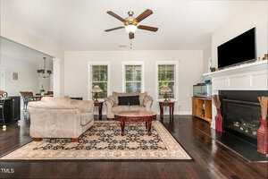 A living room with two beige sofas, a patterned area rug, and a wooden coffee table. There are three windows on the back wall, a fireplace with a TV above it, a ceiling fan, and dark hardwood floors.